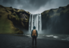 The Best Countries for Solo Travelers in 2023 a lone figure stands facing a majestic waterfall, back to the camera, wearing a weathered backpack, dark storm clouds gathering overhead