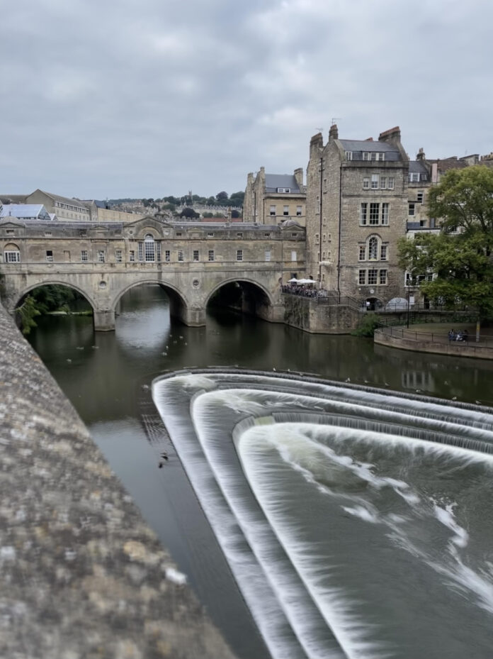 Pulteney Bridge, A stone bridge with three arches and shops on both sides, spanning over a river with a waterfall. The bridge has a classical style with pediments, pilasters, and domes. The bridge is surrounded by trees and buildings in the background