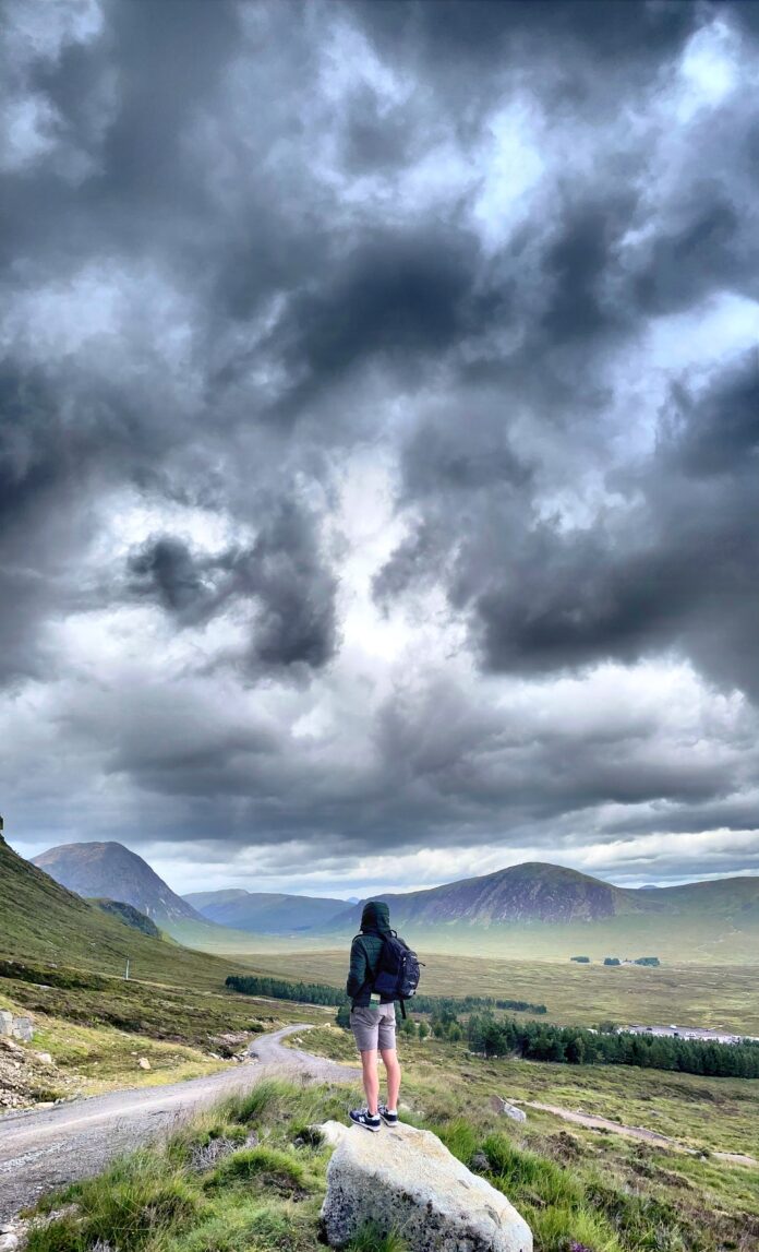 A hiker over 40 stands proudly on a Highland trail, looking out over a vast valley with rugged peaks and heather-covered moorland.